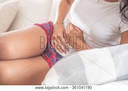 Detail Of A Young Female Sitting On Sofa, Leaning To One Side And Holding Her Aching Stomach With Bo