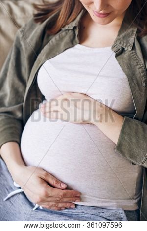 Close-up of young pregnant woman in casual clothing embracing belly while thinking about baby in her womb