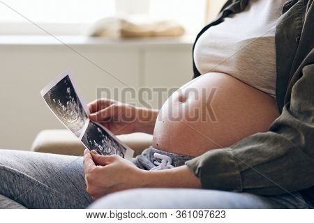 Close-up of unrecognizable pregnant woman looking at fetal ultrasound image while thinking of baby in her womb