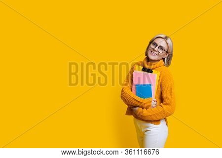 Blonde Student With Eyeglasses Smiling At Camera And Holding Some Books While Posing On A Yellow Wal