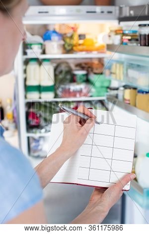Woman Standing In Front Of Refrigerator In Kitchen With Notebook Writing Weekly Meal Plan