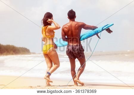 Back Side Of Young Man Holding Serf Board In Hand And Woman Wearing Yellow Swimsuit Walking On The B