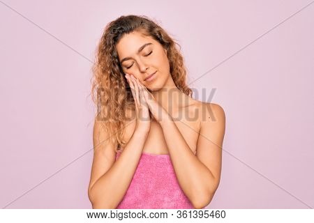 Beautiful blonde woman with blue eyes wearing towel shower after bath over pink background sleeping tired dreaming and posing with hands together while smiling with closed eyes.