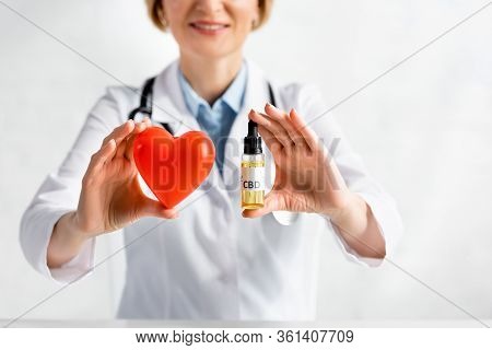 Cropped View Of Cheerful And Mature Doctor Holding Bottle With Cbd Lettering And Red Heart In Clinic