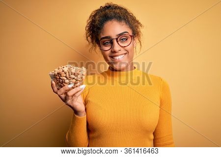 Young african american afro girl holding bowl with healthy peanuts over yellow background with a happy face standing and smiling with a confident smile showing teeth