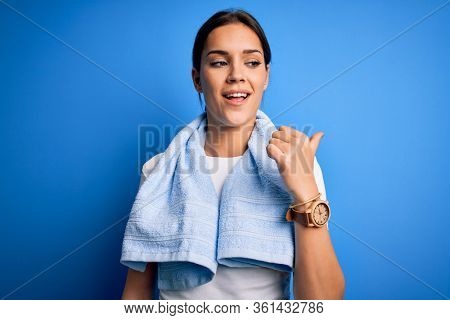 Young beautiful brunette sportswoman wearing towel doing exercise over blue background smiling with happy face looking and pointing to the side with thumb up.