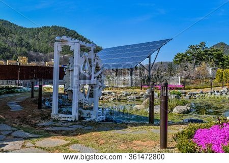 White Wooded Waterwheel In Front Of Pond In Public Park With Solar Panels In Background.
