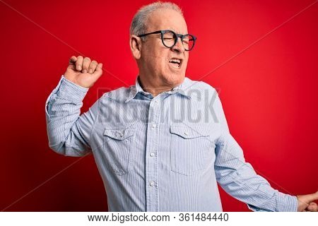 Middle age handsome hoary man wearing casual striped shirt and glasses over red background Dancing happy and cheerful, smiling moving casual and confident listening to music