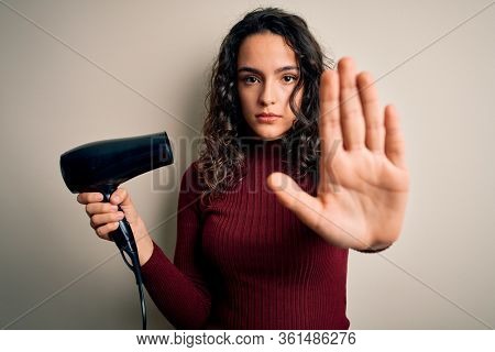 Young beautiful woman with curly hair using hair dryer over isolated white background with open hand doing stop sign with serious and confident expression, defense gesture