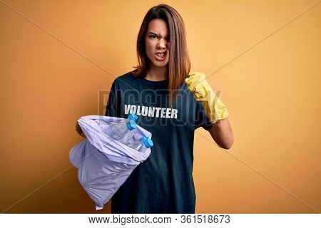 Beautiful volunteer girl caring environment doing volunteering holding bag with rubish bottles annoyed and frustrated shouting with anger, crazy and yelling with raised hand, anger concept