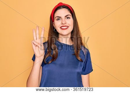 Young beautiful brunette woman wearing red lips over yellow background showing and pointing up with fingers number three while smiling confident and happy.