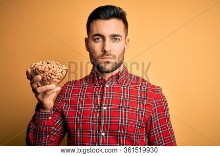 Young handsome man holding bowl with healthy peanuts over isolated yellow background with a confident expression on smart face thinking serious