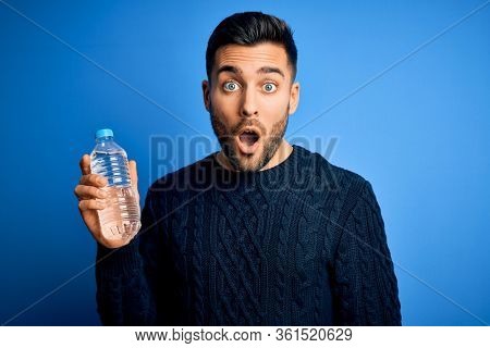 Young handsome man drinking bottle of water to refeshment over blue background scared in shock with a surprise face, afraid and excited with fear expression