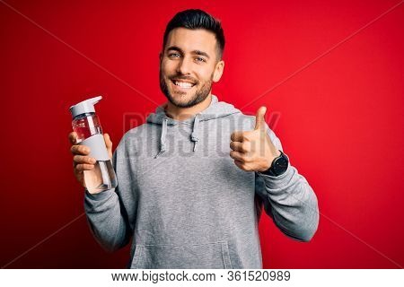 Young handsome sportsman drinking bottle of water to refeshment over red background happy with big smile doing ok sign, thumb up with fingers, excellent sign
