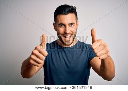 Young handsome man wearing casual t-shirt standing over isolated white background approving doing positive gesture with hand, thumbs up smiling and happy for success. Winner gesture.