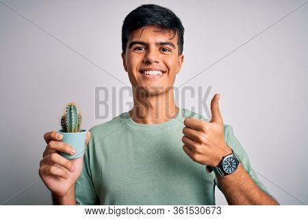 Young handsome man holding small cactus plant pot over isolated white background happy with big smile doing ok sign, thumb up with fingers, excellent sign