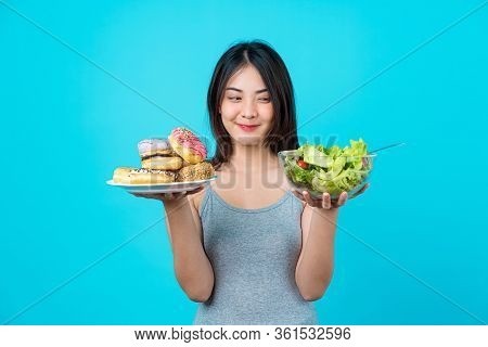 Attractive Asian Young Woman Holding And Choosing Between Disk Of Donuts Or Vegetable Salad In Glass