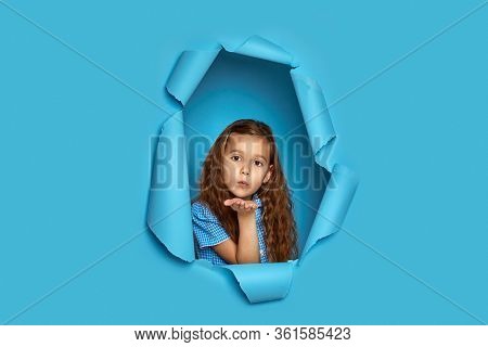 Portrait Of Cute Happy Smiling Curly Caucasian Little Cild Girl Sending Kiss In Blue Paper Hole.
