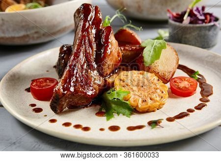 Baked ribs with homemade potatoes and coleslaw salad in white plate. Tasty meat appetizer with greenery leaves and tomatoes decorations. Delicious snack, served restaurant meal, european cuisine food