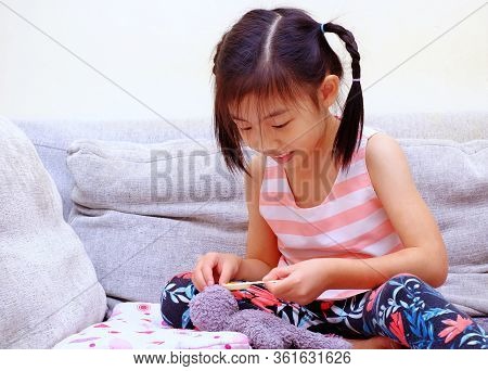 A Cute Asian Girl Playing Nurse With Her Teddy Bear. Feeding Medicine With A Small Spoon.