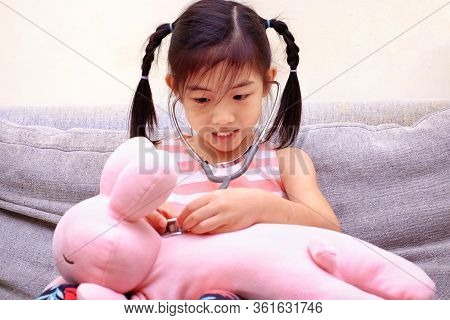 An Asian Girl Playing Doctor With A Stethoscope On Her Pink Rabbit, Smiling.
