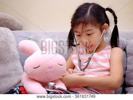 An Asian Girl Playing Doctor With A Stethoscope On Her Pink Rabbit, Smiling.