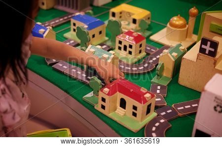 A Asian Girl Adding Wooden Road Blocks To Her Modeled Town With Buildings, School, Church And Hospit