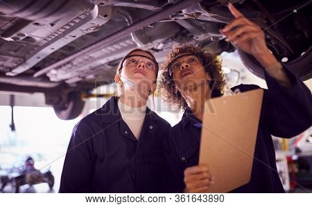 Female Tutor With Student Looking Underneath Car On Hydraulic Ramp On Auto Mechanic Course