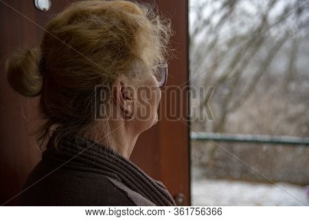 An Elderly Woman Stands In The Doorway And Looks Out At The Street.