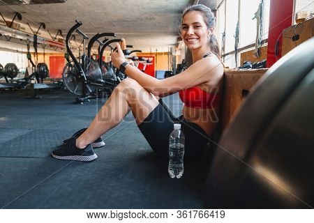 Portrait of cheerful cute sportswoman using wireless earphone and cellphone while sitting in athletic hall