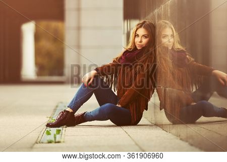 Happy young fashion woman sitting on street sidewalk  Stylish female model in brown leather fringe suede jacket and dark blue jeans