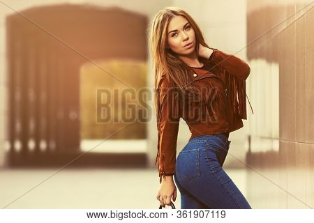 Young fashion woman with handbag walking on city street Stylish female model in brown leather fringe suede jacket and dark blue jeans
