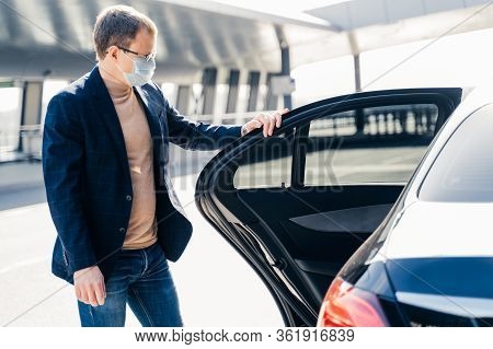 Horizontal Shot Of Man Wears Spectacles And Protective Medical Face Mask, Stands Near Car, Commutes 