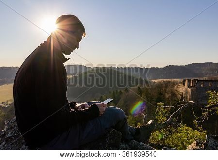 Young Caucasian Man Sitting And Using His Mobile On Rock With Ruin Divci Kamen And Sun. Czech Republ