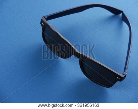 Black Square-shaped Sunglas With Blue Earhooks Against A Blue Background.