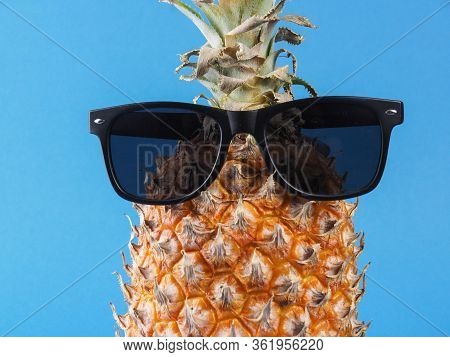 Close-up, Ripe Orange Pineapple With Green Leaves And Black Sunglas On It, On A Blue Background.