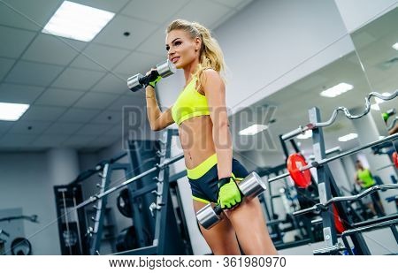 Gorgeous Blonde Woman Is Warming Up And Doing Workout At A Gym Looking At Camera. Girl Wears Bright 