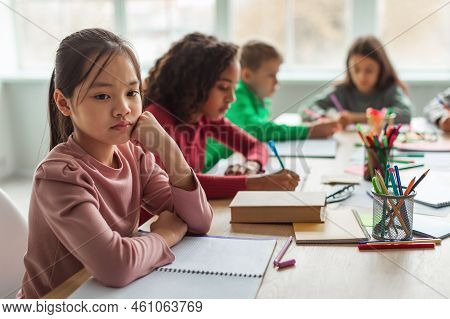 Unhappy Asian Schoolgirl Thinking Sitting With Diverse Classmates In Classroom