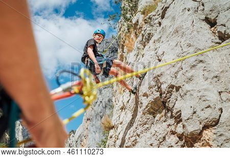 Teenager Boy On The Natural Cliff Climbing Wall. Boy Hanging On A Rope Using A Climbing Harness And