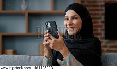 Smiling Arab Muslim Islamic 30s Woman User In Headscarf Black Hijab Using Phone At Home Sitting On C