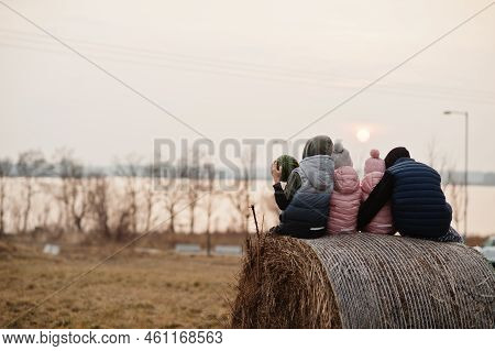 Back Of Four Kids Sitting On Haycock At Field Against Lake Sunset.
