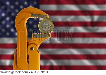 One Euro Coin Clamped In An Orange Plumber Wrench On The Background Of The Flag Of The United States