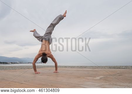 Shirtless Caucasian Man Doing Backflip On Pebble Beach.
