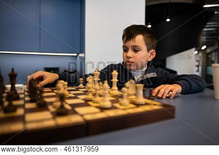 Adorable Caucasian 10 Years Old Child, Smart School Boy Sitting At A Table And Playing Chess, Though