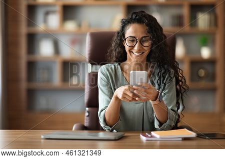 Positive Middle Eastern Woman Sitting At Workdesk, Using Smartphone