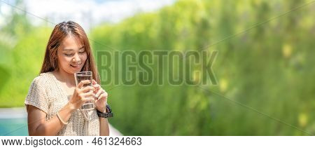 Asian Woman Holds A Glass Of Water From Drinking At Outdoor Field With Bokeh Green Banner Background