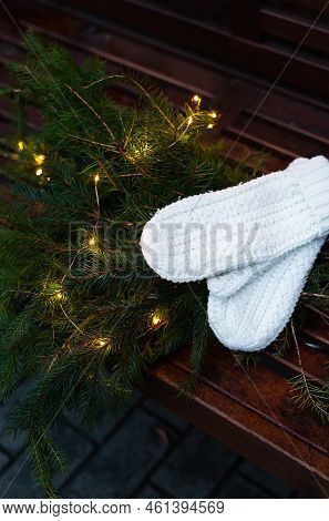 Branches Of A Coniferous Tree Decorated With A Garland Lie On A Bench Along With Knitted White Mitte