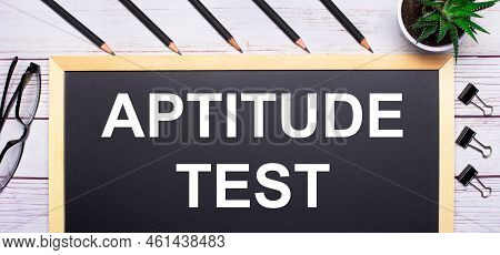 On A Light Wooden Table - A Board With The Text Aptitude Test, Pencils, Plants, Glasses And Paper Cl