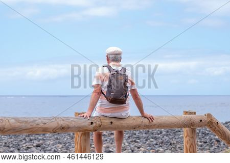 Rear View Of Relaxed Caucasian Bearded Man With Hat Sitting Near The Beach Looking At The Horizon Wh