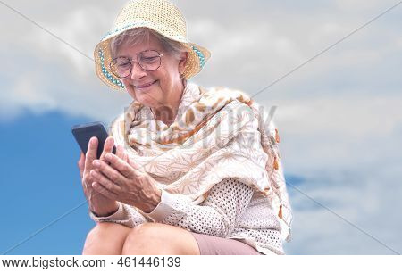 Portrait Of Smiling Senior Woman Wearing Eyeglasses And Summer Hat Using Mobile Phone Sitting Outdoo